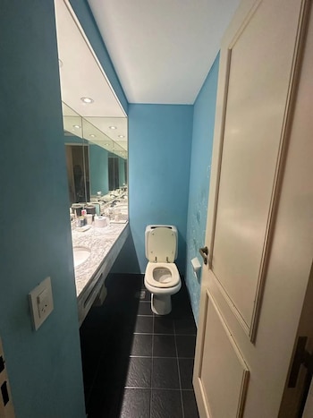 Baño pequeño con paredes azules, inodoro blanco con tapa abierta, un largo lavamanos de mármol con espejo y luces empotradas, y suelo de baldosas oscuras