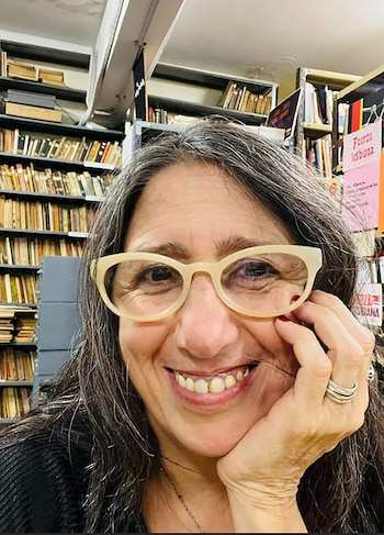 Primer plano de Daniela Azulay sonriendo, mujer de cabello gris con gafas claras y uñas rojas, apoyando la mejilla. Estanterías con libros al fondo