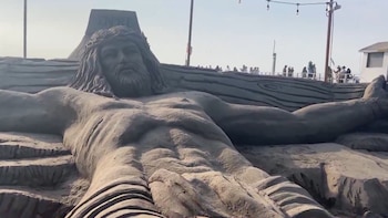 Vista cercana de una monumental escultura de arena de Jesús crucificado. Muestra su torso, brazos extendidos, cabeza con corona de espinas y barba