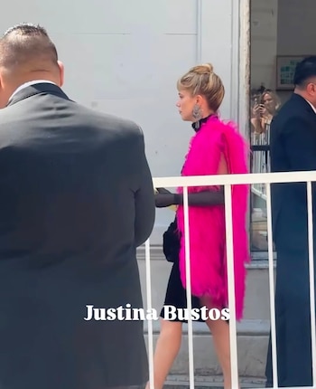 Una mujer de perfil con estola fucsia y vestido negro camina detrás de una valla blanca junto a personas vestidas con trajes oscuros