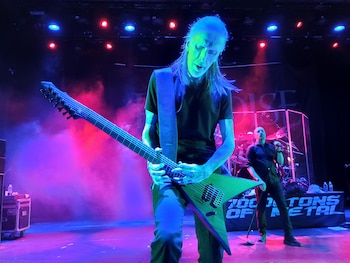 Un músico con cabello largo tocando una guitarra eléctrica en forma de V con múltiples cuerdas, iluminado por luces rojas y azules en un escenario con humo