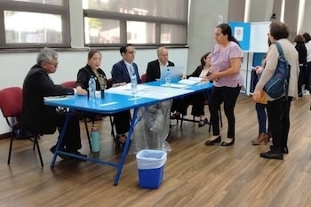 Cinco personas sentadas en una mesa azul con documentos y botellas de agua. Tres personas de pie conversan. Un cesto azul con bolsa blanca está en el suelo