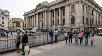Peatones en una acera junto a una reja metálica, con una avenida con autobuses y un edificio institucional de columnas de piedra al fondo.