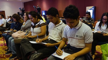 Jóvenes y adultos sentados en filas dentro de un auditorio escriben en hojas de papel sobre carpetas.