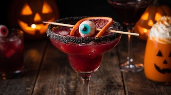 Copa de margarita con bebida roja, escarchado negro, una rodaja de naranja sanguina y un ojo de gomita atravesado por un palillo sobre madera.