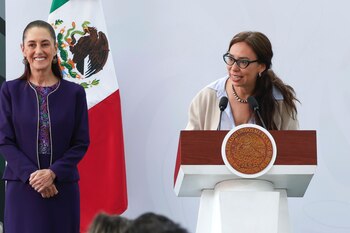 Claudia Sheinbaum viste traje morado junto a la bandera mexicana mientras una mujer con anteojos habla frente a un pódium
