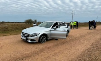 Mercedes Benz plateado con puertas abiertas y rueda delantera izquierda dañada en camino de tierra. Agentes con un perro están detrás en un campo con cielo nublado
