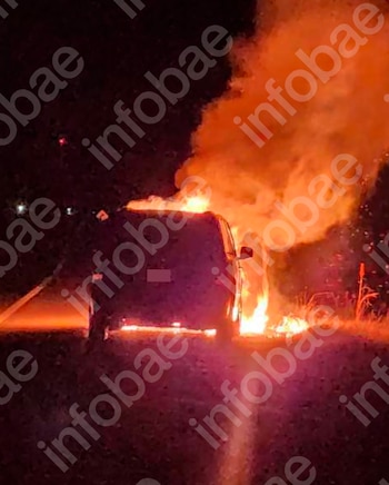 Un automóvil de color oscuro arde en llamas intensas en el costado de una ruta durante la noche. Las llamas son grandes y emiten humo visible