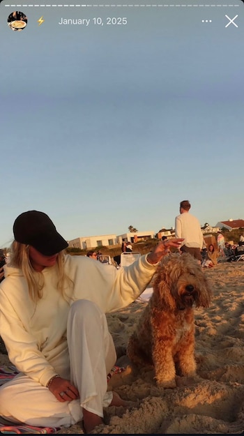 Mujer rubia con gorra negra y vestimenta clara, sentada en la arena, acaricia a un perro de pelo rizado. Fondo con personas, edificios y cielo claro