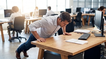 Hombre de camisa y pantalón oscuro hace flexiones sobre un escritorio de madera. Computadoras, teclados, papeles y empleados al fondo.