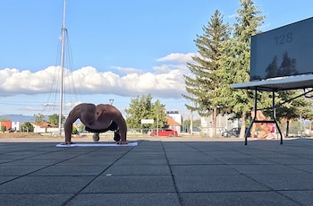 Un hombre hace flexiones en el suelo junto a una mesa con una pantalla de televisión, con pinos y vehículos de fondo