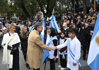 Un hombre de saco claro estrecha la mano de un niño. Varias personas y jóvenes con banderas argentinas. Fondo con edificios, árboles y un semáforo
