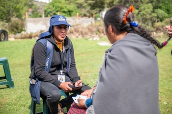 Dos mujeres sentadas en taburetes en un área verde. Una lleva gorra azul con el logo 'RSH' y mochila, y sostiene un teléfono. La otra tiene trenzas y sujeta tarjetas