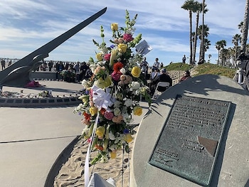 Una placa de bronce grabada sobre piedra junto a un arreglo floral de colores en una plaza costera con personas y palmeras al fondo