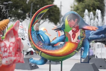 Escultura artística de colores vivos con la pintura de un hombre y un ave, sobre una base negra. Otras esculturas y una fuente borrosa en el fondo
