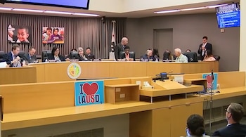 Sala de reuniones con mesa curva y personas sentadas. Pantalla con logo del Distrito Escolar Unificado de Los Ángeles. Letrero 'I Love LAUSD'. Pantallas con niños