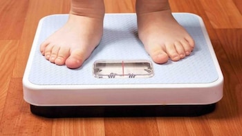 Pies descalzos de un niño o niña sobre una balanza doméstica de color blanco y azul. El suelo es de madera