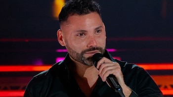 Carlos Marchesi, un hombre con barba y pelo oscuro, sostiene un micrófono con expresión emotiva y ojos llorosos. El fondo es oscuro con luces rojas y púrpuras