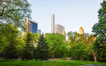 Vista de un parque verde con árboles frondosos y senderos, enmarcando el horizonte de Nueva York con rascacielos como 432 Park Avenue bajo un cielo claro