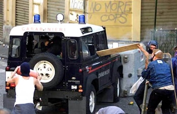 Un vehículo oscuro de Carabinieri con luces azules es atacado por tres personas. Una sujeta una tabla, otra un cilindro rojo. Graffiti "NO MORE COPS"