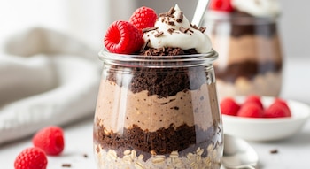 Un tarro de cristal con avena trasnochada de pastel de chocolate, capas de avena, crema, migas de pastel, frambuesas y crema batida con virutas de chocolate.