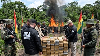 Agentes de la DEA y militares bolivianos uniformados junto a una gran pila de paquetes de drogas, con barriles en llamas y humo negro en un entorno selvático.