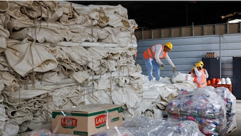 Vista interior de un almacén donde trabajadores con chalecos y cascos de seguridad organizan grandes pilas de sacos de lona blanca y cajas de ayuda humanitaria