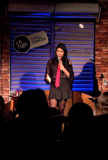 Mujer en escenario con camisa negra, falda, corbata roja, medias, y un micrófono. Fondo con muro de ladrillo, persiana, y letrero Club de Comedia