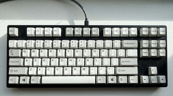 Vista superior de un teclado de computadora mecánico con teclas blancas cuadradas y una base negra, conectado por un cable trenzado gris.
