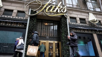 La fachada de la tienda Saks Fifth Avenue en la ciudad, con su gran letrero dorado, entrada decorada con follaje y personas caminando por la acera