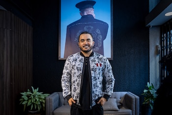 Hombre sonriente con barba y chaqueta estampada, manos en los bolsillos, frente a una gran fotografía y un sofá gris