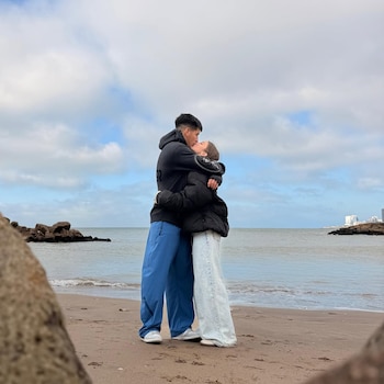 Pareja joven besándose en una playa con el mar de fondo. Él viste sudadera negra y pantalón azul, ella chaqueta negra y pantalón claro. Cielo nublado