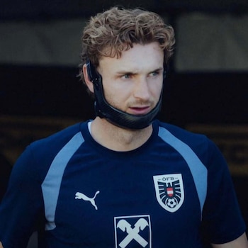 Primer plano de Stefan Posch con cabello castaño ondulado, camiseta azul con logos de Puma y ÖFB, y protector facial negro en la barbilla