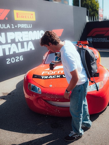 Un hombre con camiseta blanca vierte agua frente a un auto rojo con ojos y boca dibujados, junto a un cartel publicitario