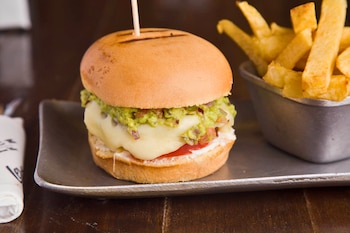 Hamburguesa con pan tostado, guacamole, queso derretido y tomate, servida en una bandeja metálica con un vaso de papas fritas a la derecha
