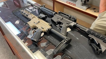 Dos rifles, uno de color beige y otro negro, sobre un mostrador de cristal con un tapete que dice 'AR-15 CARBINE RIFLE' en una tienda