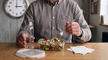 Hombre sentado con camisa gris, contenedor de ensalada con tenedor, botella de agua, servilletas, tapa de plástico y reloj de pared.