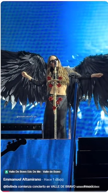 Una mujer vestida con alas negras de plumas y gafas de sol canta ante un pedestal de micrófono decorado con flores