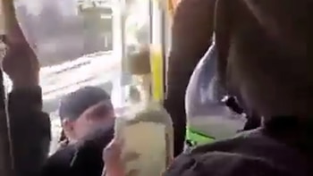 Interior de un colectivo con varias personas; una mano sostiene una botella de vidrio con una etiqueta amarilla; otra botella es visible con un detalle verde