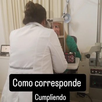 Una médica con bata blanca examina los ojos de una mujer rubia con chaqueta verde mediante un equipo oftalmológico