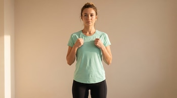 Mujer de cabello recogido, camiseta verde menta y leggings negros posa con puños al pecho sobre fondo beige. Iluminación cálida lateral.