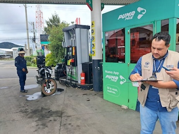 Un hombre con casco de seguridad naranja y chaleco sostiene una tabla con hojas en la pista de una gasolinera con surtidores