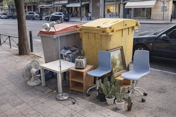 Objetos voluminosos desechados como muebles, electrodomésticos y plantas junto a contenedores de basura en una acera urbana con coches y edificios.