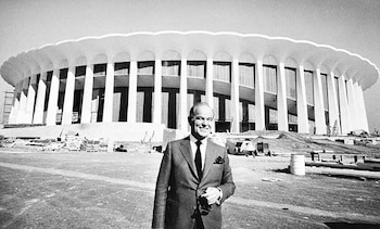 Fotografía en blanco y negro de un hombre con traje sonriendo frente a un gran estadio circular en construcción con columnas blancas