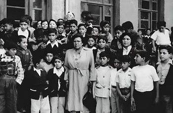 Grupo de niños y dos mujeres adultas, una con gafas y un abrigo con cinturón, frente a un edificio con ventanas. Imagen en blanco y negro