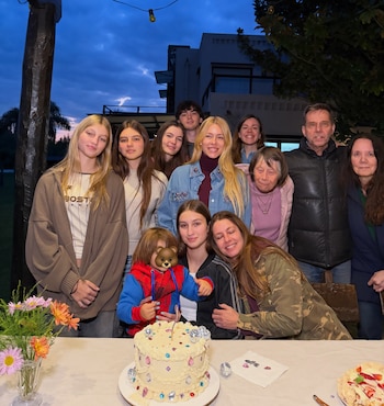 Un grupo de personas posa al aire libre detrás de una mesa con una torta decorada y un florero frente a una vivienda