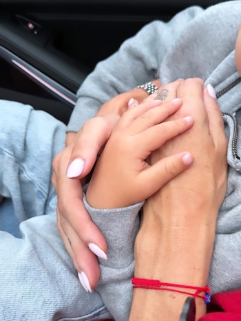 Primer plano de una mano adulta con uñas claras y un anillo, sosteniendo la pequeña mano de un niño. Se ven detalles de ropa gris y un brazalete rojo