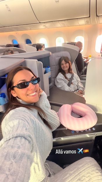 Mujer con gafas de sol y niña sonríen en cabina de clase ejecutiva de avión. Almohada de cuello rosa. Texto 'Allá vamos', avión y bandera de España