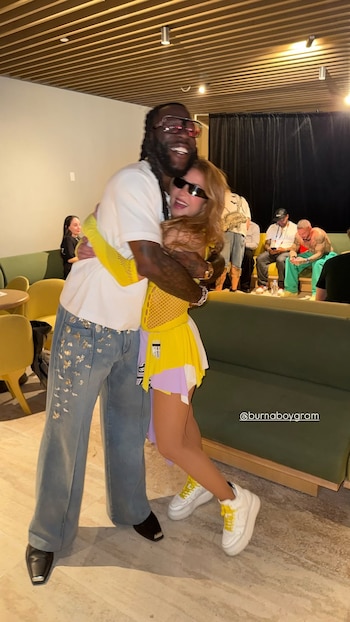 Shakira, con cabello claro, top amarillo, falda y gafas de sol, abraza a Burna Boy, con rastas, camiseta blanca, jeans y gafas oscuras, ambos sonríen
