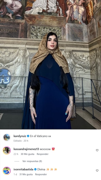 Karely Ruiz de pie dentro de un edificio con elaborados murales y relieves. Viste un vestido azul oscuro y una pashmina dorada cubriendo su cabeza